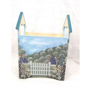 May include: A wooden storage box with a painted scene of a garden with a white picket fence. The box is painted in shades of blue, green, and white, with a yellow top.