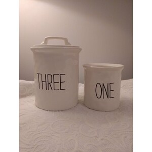May include: Two white ceramic canisters with black lettering. The larger canister says "THREE" and the smaller canister says "ONE".