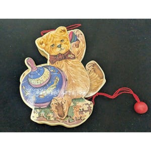 May include: A wooden pull toy featuring a brown teddy bear holding a blue and gold spinning top. The bear is wearing a red bow tie and has a red string attached to its hand. The toy is on a white background.
