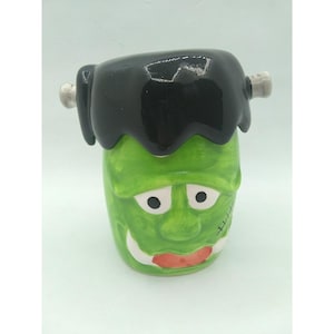 Peut inclure: Une tasse en céramique verte représentant le monstre de Frankenstein, avec des cheveux noirs, des boulons blancs et une cicatrice cousue. La tasse a une expression souriante avec une bouche rouge et des dents blanches. Un objet de collection sur le thème d'Halloween.