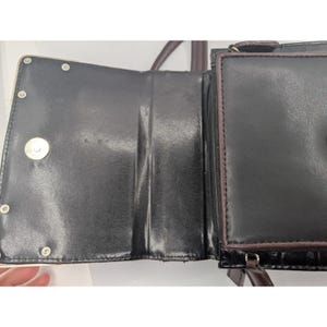 K&ouml;nnte beinhalten: Offene schwarze Ledergeldb&ouml;rse mit mehreren F&auml;chern. Die Geldb&ouml;rse hat einen Magnetverschluss und eine Rei&szlig;verschlusstasche. Die Innenseite ist schwarz mit gl&auml;nzendem Finish. Die Geldb&ouml;rse hat einen braunen Riemen.