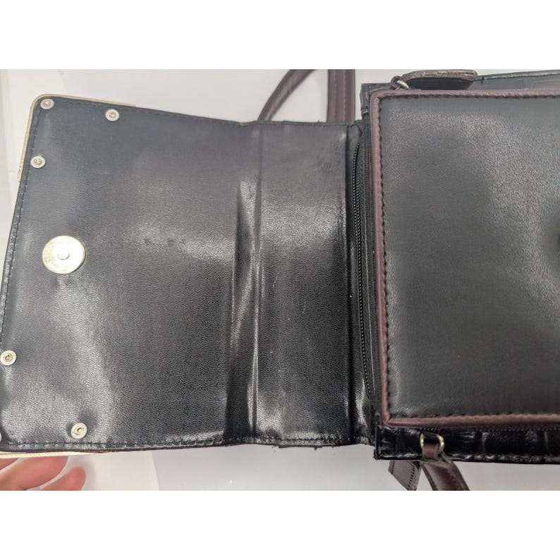 K&ouml;nnte beinhalten: Offene schwarze Ledergeldb&ouml;rse mit mehreren F&auml;chern. Die Geldb&ouml;rse hat einen Magnetverschluss und eine Rei&szlig;verschlusstasche. Die Innenseite ist schwarz mit gl&auml;nzendem Finish. Die Geldb&ouml;rse hat einen braunen Riemen.