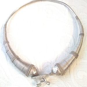 May include: A silver choker necklace with a unique design. The necklace is made of multiple thin wires that are twisted together to create a textured look. The ends of the necklace are rounded and have a decorative design.