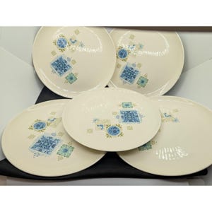 Peut inclure: Un ensemble de cinq assiettes en céramique blanc cassé. Chaque assiette présente un motif répétitif de formes géométriques bleues et vertes et des accents floraux. Les assiettes sont disposées sur une surface noire.