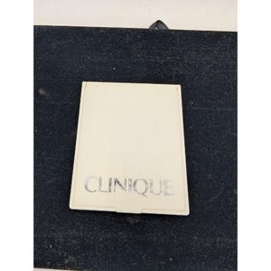May include: A rectangular, off-white compact mirror featuring the brand name CLINIQUE printed in silver lettering on the front cover. The compact has a smooth, flat surface and a hinged design, presenting a clean and functional aesthetic.