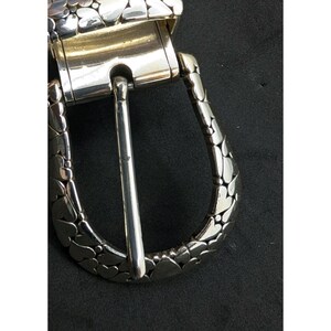 May include: Close-up of a silver-toned belt buckle. The buckle features a rectangular frame with a pivoting bar and a heart-shaped pattern on the outer edges. The pin is a simple, polished silver. The background is black.