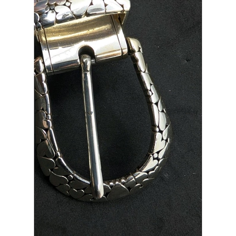 May include: Close-up of a silver-toned belt buckle. The buckle features a rectangular frame with a pivoting bar and a heart-shaped pattern on the outer edges. The pin is a simple, polished silver. The background is black.