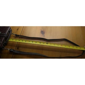 K&ouml;nnte beinhalten: Eine braune Leder-Umh&auml;ngetasche mit strukturiertem Muster. Die Tasche hat einen langen, verstellbaren Riemen. Ein gelbes Ma&szlig;band zeigt, dass der Riemen etwa 56 cm lang ist.