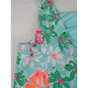 May include: A sleeveless dress with a floral pattern. The dress features a light blue background with large flowers in shades of green, white, pink, and orange. The fabric has a textured appearance.