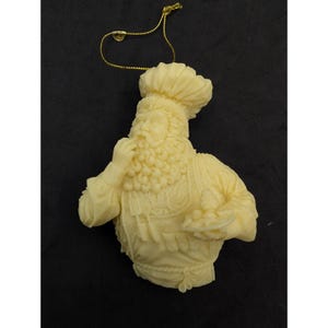 Op de afbeelding: Een crèmekleurig, gegoten ornament dat een chef-kok met een hoge muts en schort voorstelt. Het figuur heeft een krullende baard, houdt een klein bordje met eten in de ene hand en raakt met de andere hand de kin aan. Een gouden koord is aan de bovenkant bevestigd.