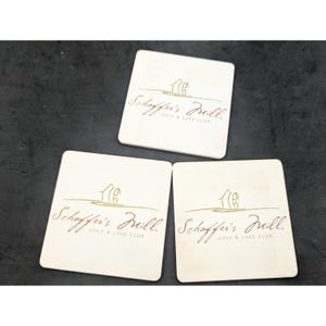 May include: Three white square coasters with a brown logo that says "Schaffer's Mill Golf & Lake Club". The logo features a stylized drawing of a house with a windmill.