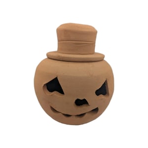 May include: A light brown, unglazed terracotta jack-o'-lantern figurine with a rounded body and a top hat. The pumpkin features carved, angular eye sockets, a small triangular nose, and a wide, jagged mouth opening, revealing a dark interior.