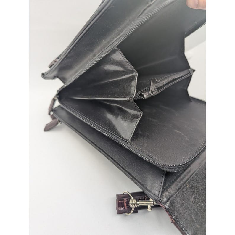 K&ouml;nnte beinhalten: Offene schwarze Ledergeldb&ouml;rse mit mehreren F&auml;chern und einer Rei&szlig;verschlusstasche. Die Geldb&ouml;rse hat eine braune Handschlaufe mit einem silbernen Verschluss. Der Innenraum ist schwarz mit gl&auml;nzendem Finish.