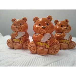 May include: Three brown ceramic bear figurines. Each bear is seated, holding a white object and a honey pot. The bears have black eyes and are smiling. The honey pots are yellow and brown.