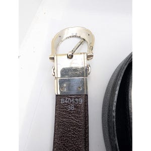 May include: A brown leather belt with a silver-colored buckle. The belt is marked with the numbers "B40539" and "38". The buckle has a decorative, curved design. The belt is partially visible against a white background.