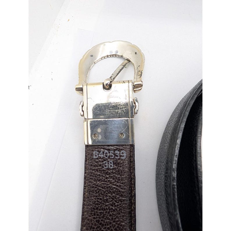 May include: A brown leather belt with a silver-colored buckle. The belt is marked with the numbers "B40539" and "38". The buckle has a decorative, curved design. The belt is partially visible against a white background.
