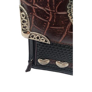 K&ouml;nnte beinhalten: Eine braune und schwarze Geldb&ouml;rse mit einem strukturierten, krokodilartigen Muster. Die Geldb&ouml;rse hat silberfarbene dekorative Metallakzente an den Ecken und an der Vorderseite sowie einen gewebten schwarzen Abschnitt und einen Rei&szlig;verschluss.
