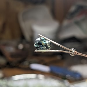 May include: A round, faceted gemstone with a green and blue color held by tweezers.