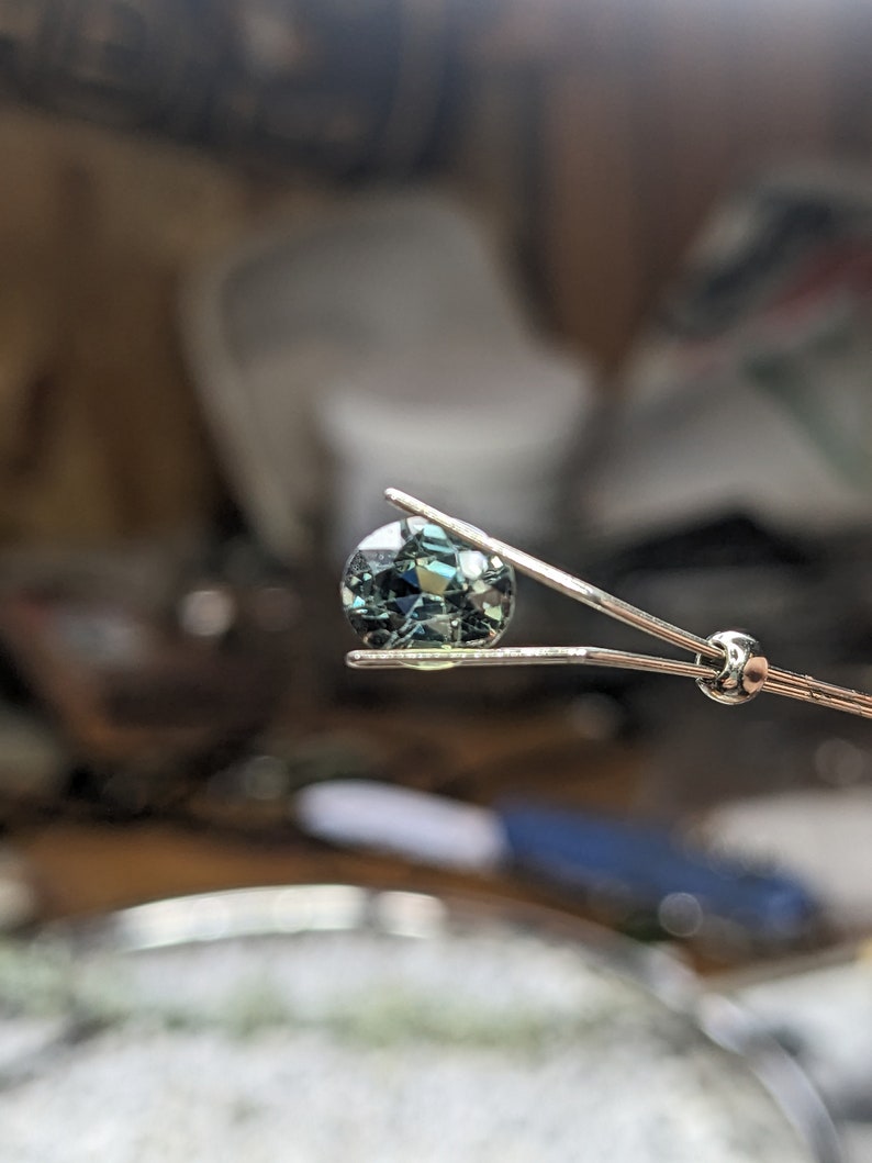 May include: A round, faceted gemstone with a green and blue color held by tweezers.