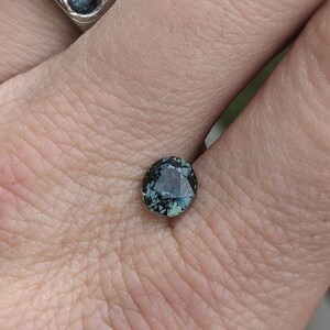 May include: A dark green gemstone with a faceted cut. The gemstone is set in a ring and is being held by a hand.