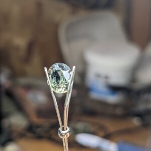 May include: A green gemstone is held in a metal prong setting. The gemstone is oval shaped and has a faceted cut.