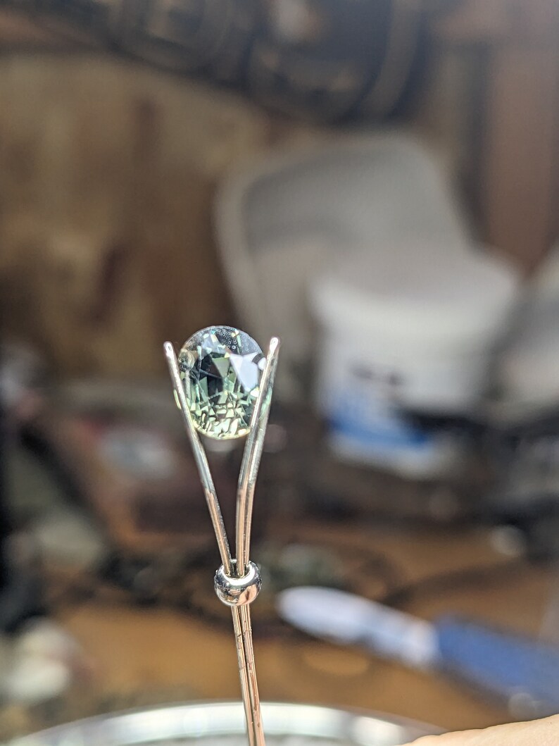 May include: A green gemstone is held in a metal prong setting. The gemstone is oval shaped and has a faceted cut.