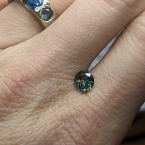 May include: A round, green gemstone with a dark blue hue. The gemstone is faceted and has a high polish.