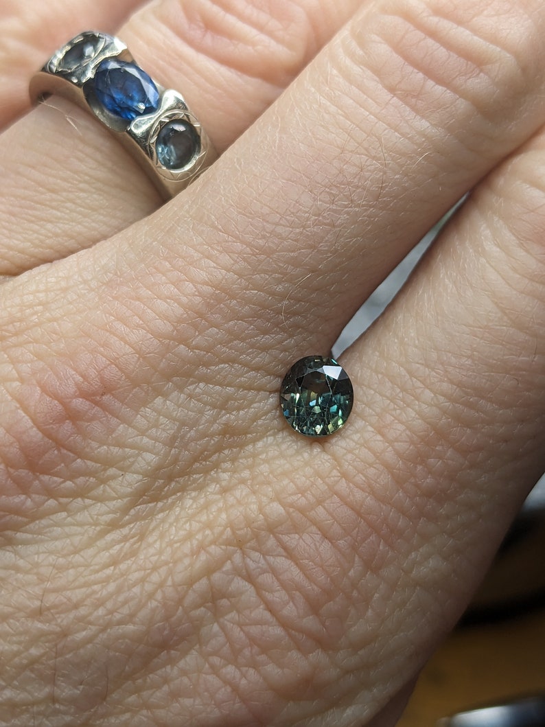 May include: A round, green gemstone with a dark blue hue. The gemstone is faceted and has a high polish.