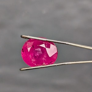 May include: A faceted, oval ruby gemstone held by tweezers. The ruby is a vibrant, deep pink colour with visible internal inclusions. The tweezers are silver-coloured, and the background is a solid grey.