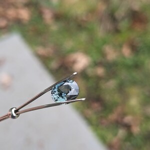May include: A teal blue oval gemstone held in tweezers against a blurred background.