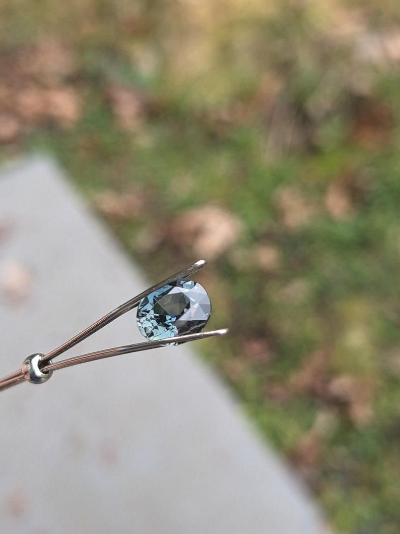 May include: A teal blue oval gemstone held in tweezers against a blurred background.