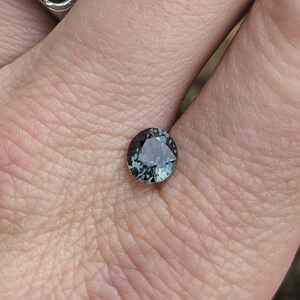 May include: A round, dark green gemstone with a slight blue hue. The gemstone is set in a ring and is being held by a hand.