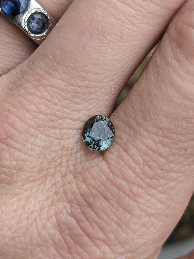May include: A round, dark green gemstone with a slight blue hue. The gemstone is set in a ring and is being held by a hand.
