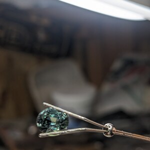 May include: A teal gemstone held in a pair of silver tweezers.