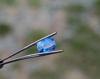 May include: A close-up of a light blue oval gemstone held by tweezers.