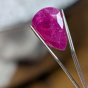 Puede incluir: Una gema de rubí facetada en forma de pera, sujeta con pinzas plateadas. El rubí es de un color rojo rosado intenso y vibrante, con inclusiones internas visibles y una superficie pulida. Las pinzas están enfocadas.