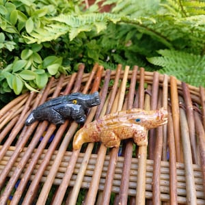 May include: Two carved alligator figurines, one is black and the other is brown with red and white markings. The figurines are sitting on a woven wicker surface.