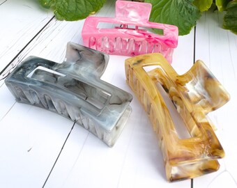 May include: Three hair clips in different colours and patterns. The clips are made of plastic and have a marbled appearance. The colours are pink, grey, and brown.