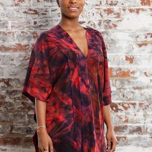 May include: A woman wearing a red, black, and white tie-dye kaftan with a v-neckline. She is standing in front of a brick wall.