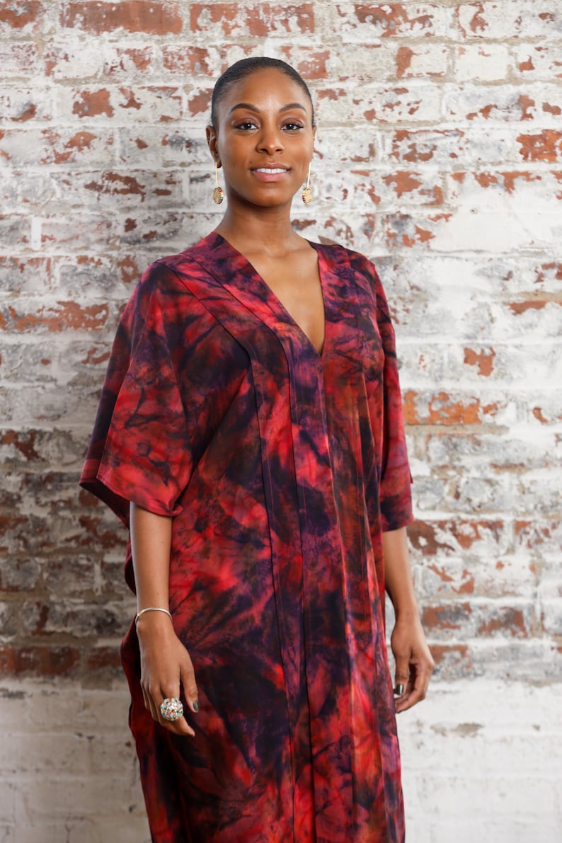 May include: A woman wearing a red, black, and white tie-dye kaftan with a v-neckline. She is standing in front of a brick wall.