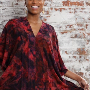 May include: A woman wearing a red and black tie-dye kaftan with a v-neck and short sleeves. The kaftan has a flowing silhouette and is made of a soft, silky fabric.