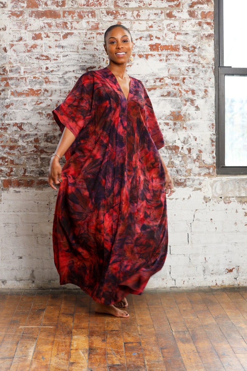 May include: A woman wearing a long, flowing red and purple tie-dye dress with a V-neckline. The dress has a loose, comfortable fit and is perfect for a casual summer day.