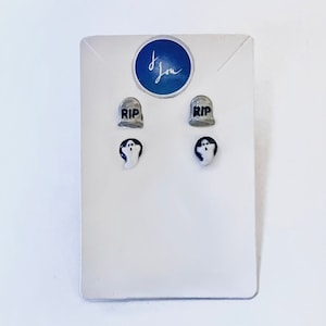 May include: A set of Halloween-themed earrings on a white card. The earrings include two pairs: gray tombstones with "RIP" and white ghost shapes with black eyes and mouths. A blue circle with white script is in the upper right corner.