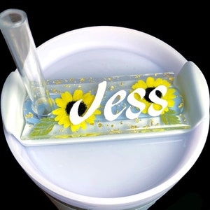 May include: White tumbler with a clear straw and a personalised nameplate with the name "Jess" in white letters and yellow sunflowers. The nameplate is embedded in clear resin with gold glitter.