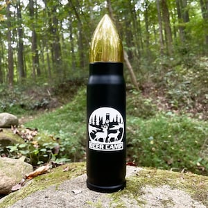 May include: Black and gold bullet-shaped thermos with a white deer camp logo featuring two deer and a tree silhouette. The logo is in a circle with the text "DEER CAMP" below it.