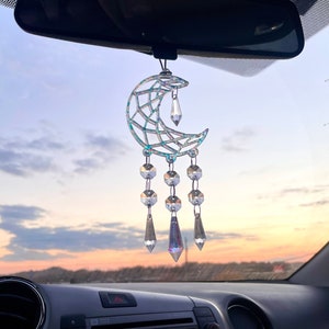 Pode incluir: Um amuleto de carro em forma de lua crescente prateada com cristais iridescentes pendurados nele. O amuleto está pendurado no espelho retrovisor de um carro.