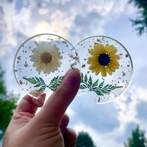 May include: Two clear resin coasters with a pressed flower design. One coaster features a white daisy with green leaves and gold flecks. The other coaster features a yellow sunflower with green leaves and gold flecks.