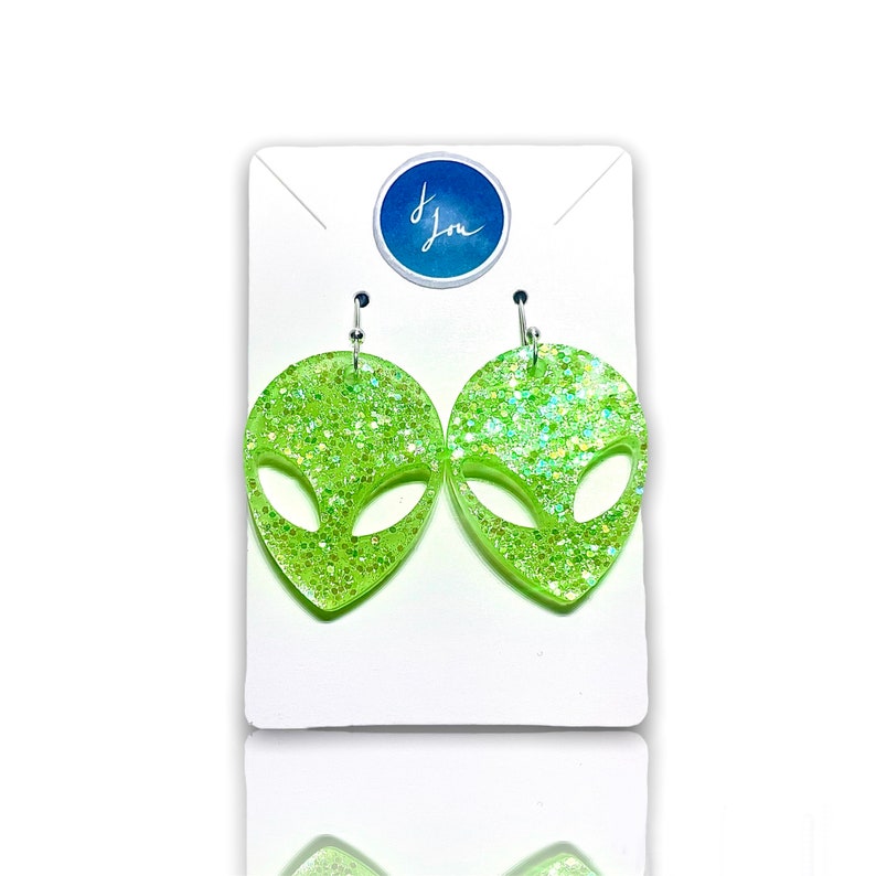 May include: A pair of green alien-shaped earrings with silver hooks. The earrings are made of clear resin with green glitter embedded in it.