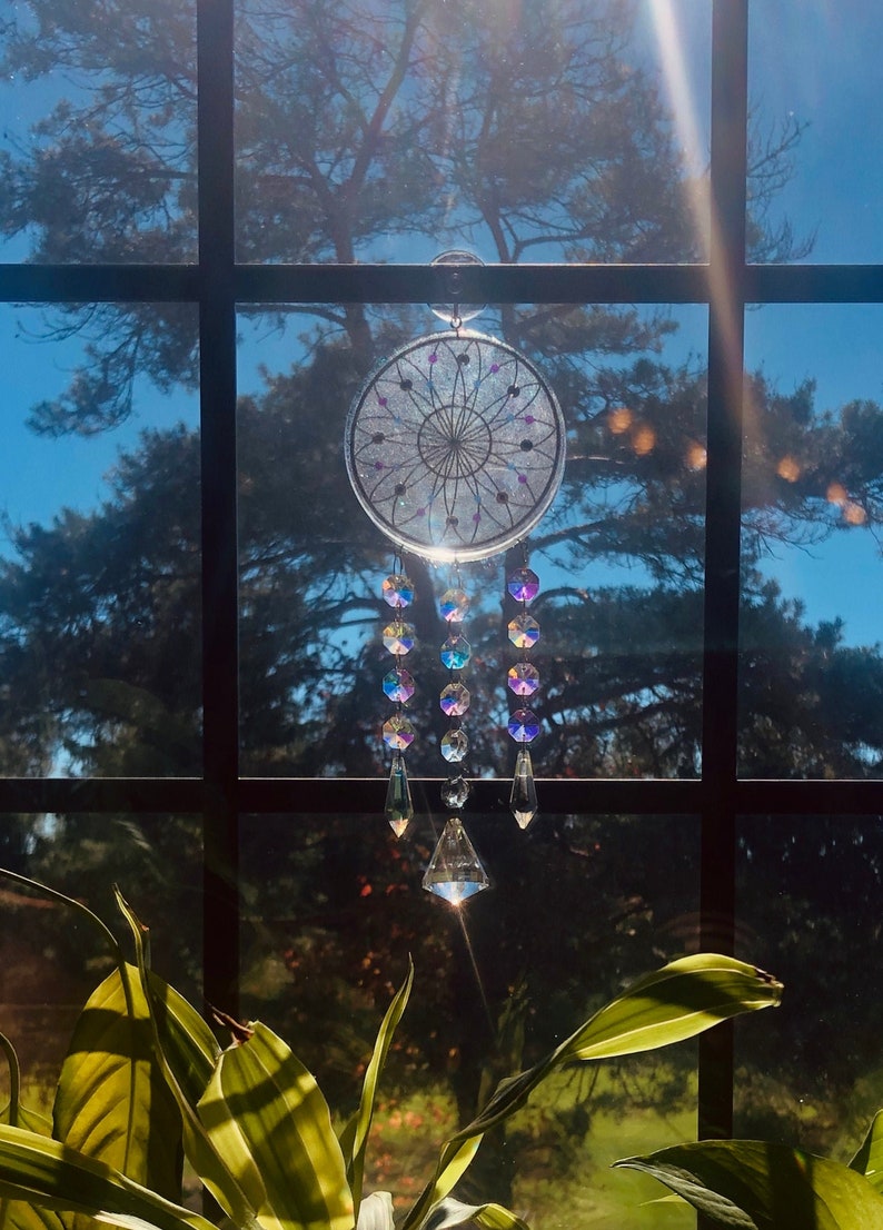 May include: A white dreamcatcher with iridescent beads and crystals hanging from it. The dreamcatcher is hanging in front of a window with a view of a tree and blue sky.