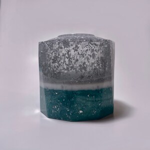 May include: A hexagonal, clear glass vase with a teal and silver glitter design. The vase has a white layer in the middle.